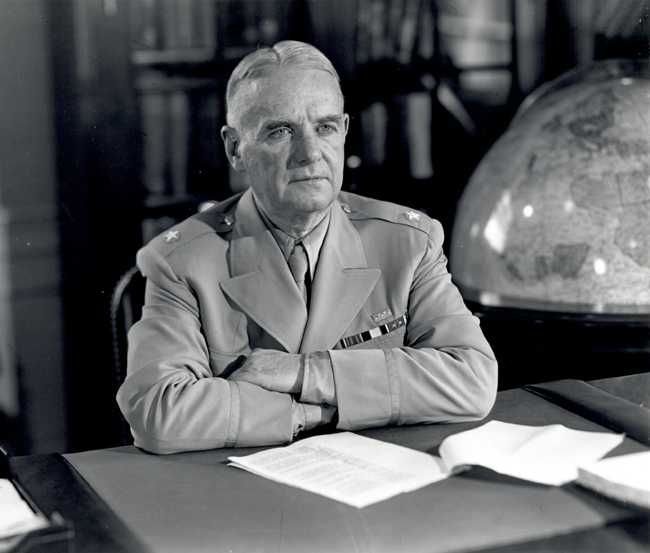 A closeup of a man in uniform at a desk with his arms folded.