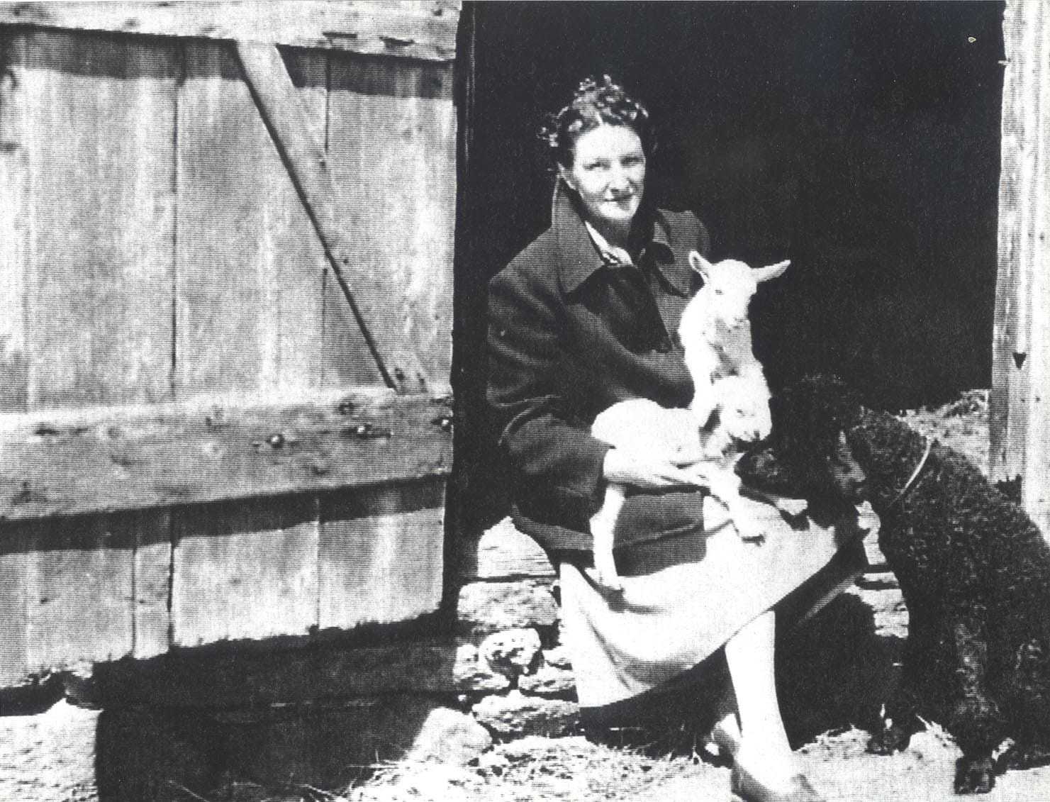 Virginia Hall holding a baby sheep, sitting next to a big black dog.