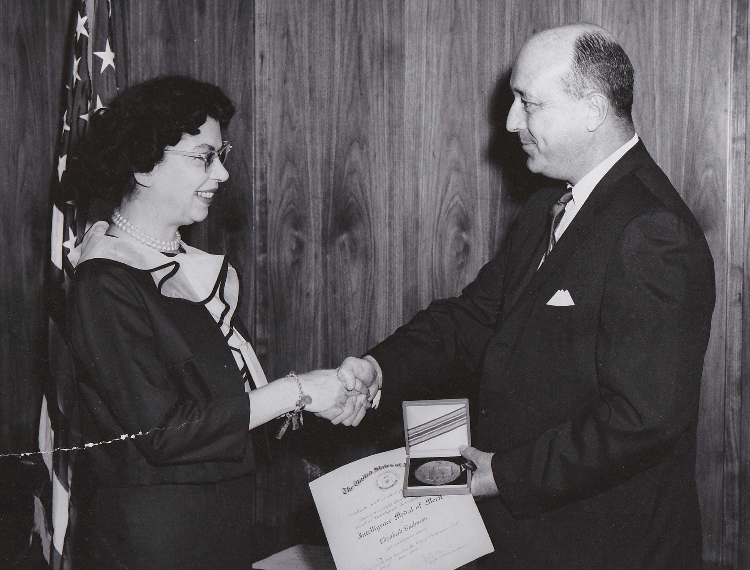 A black and white photograph of Elizabeth shaking hands in front of an American flag while receiving IMM award.