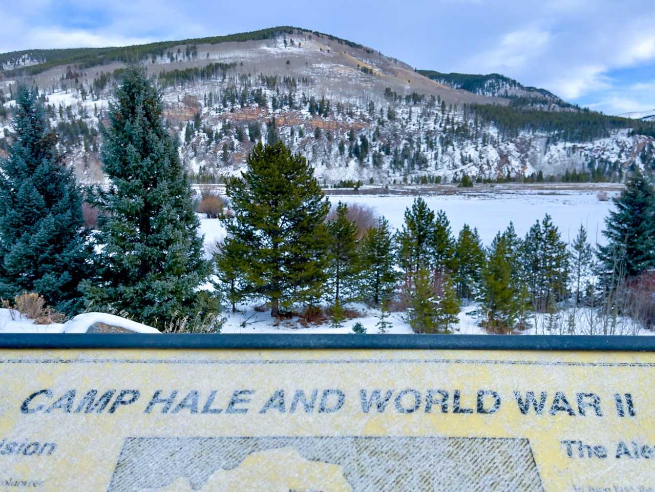 Informational sign that says Camp Hale and World War II shown at the bottom of the image, with a snowy mountain and meadow, and cascading pine trees, on the top half of the photo.