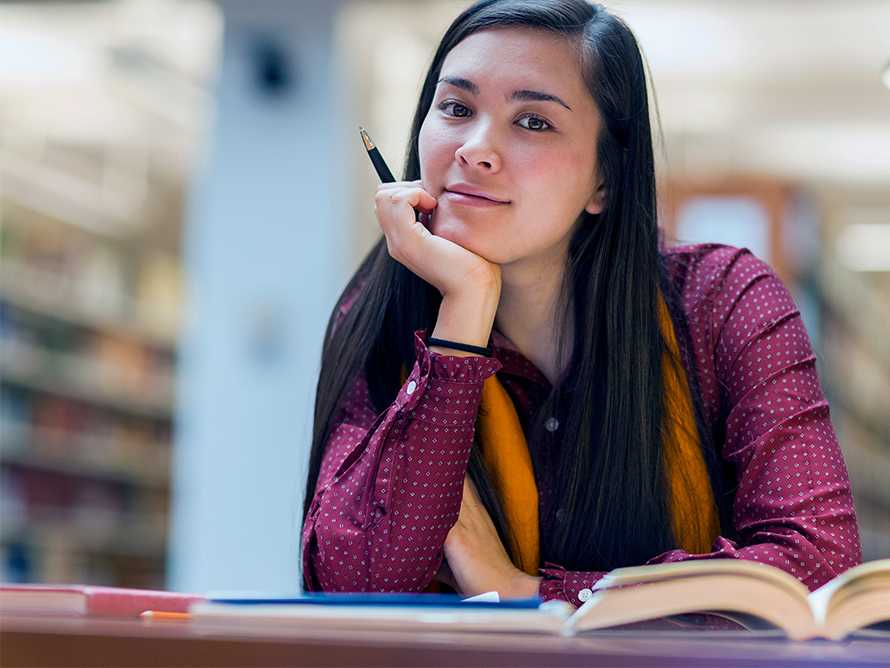 A young woman in a library holding a pen in front of a desk with several open books.