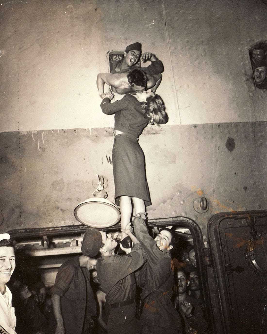 A sepia colored image of two soldiers lifting Marlene up from her feet while she kisses a soldier with his head out the window of a ship.