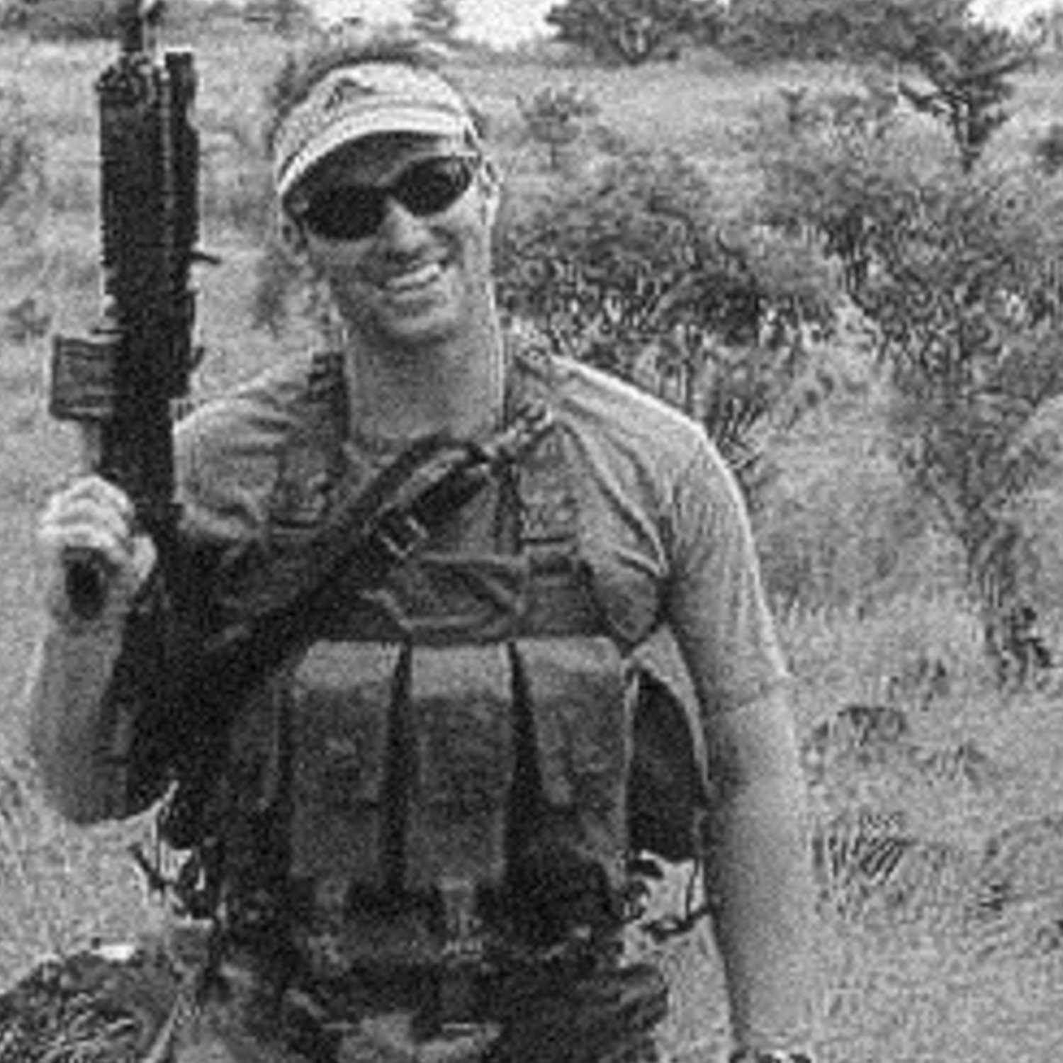 A photo of Glen A. Doherty in a military outfit and sunglasses, standing in greenery and holding a gun.