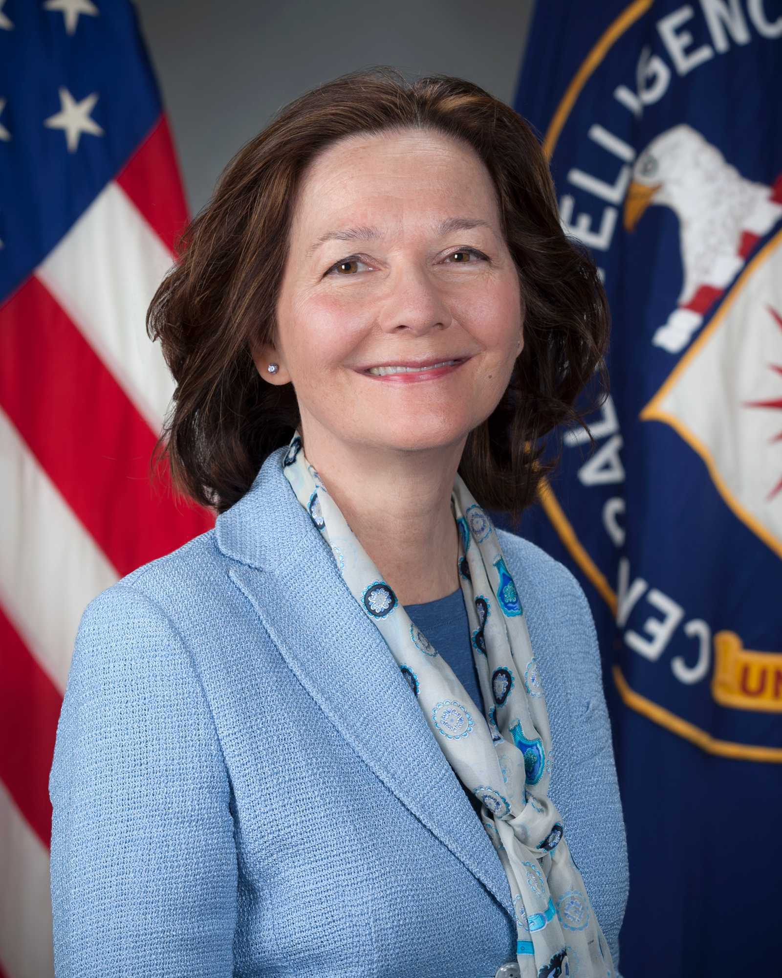 A headshot of Gina Haspel standing in front of the American flag and the CIA flag.