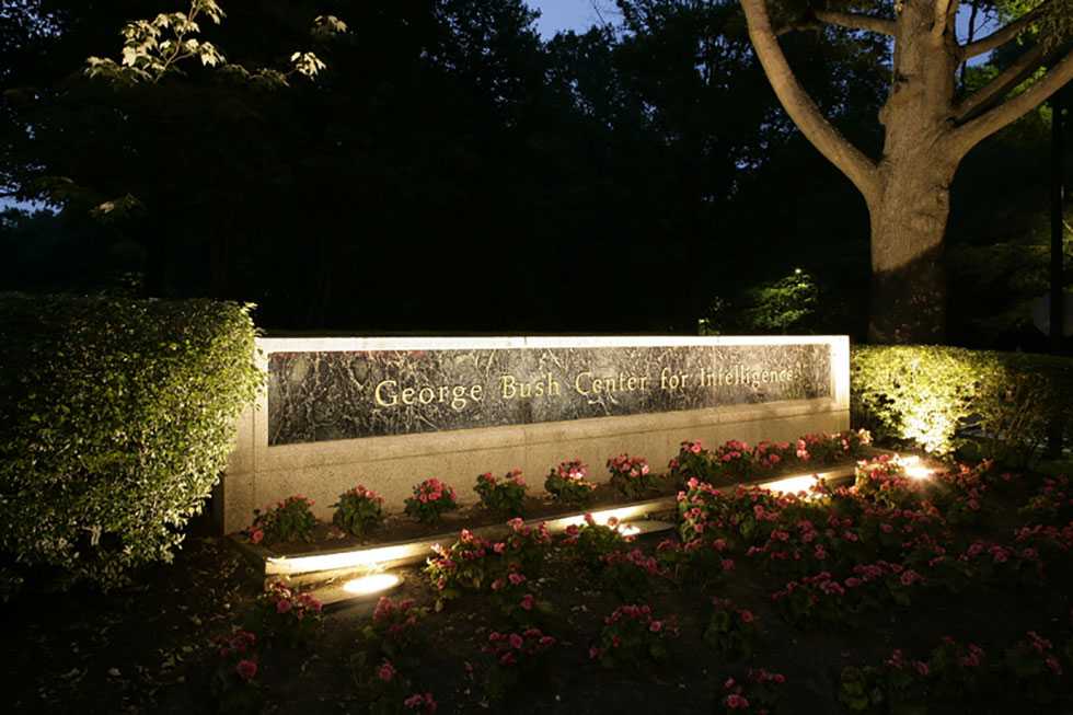 A well-lit, granite and cement wall with gold lettering, surrounded by flower beds.