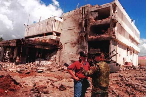 A large, pale building, on red clay, covered in ash, with an exposed side from a bombing.