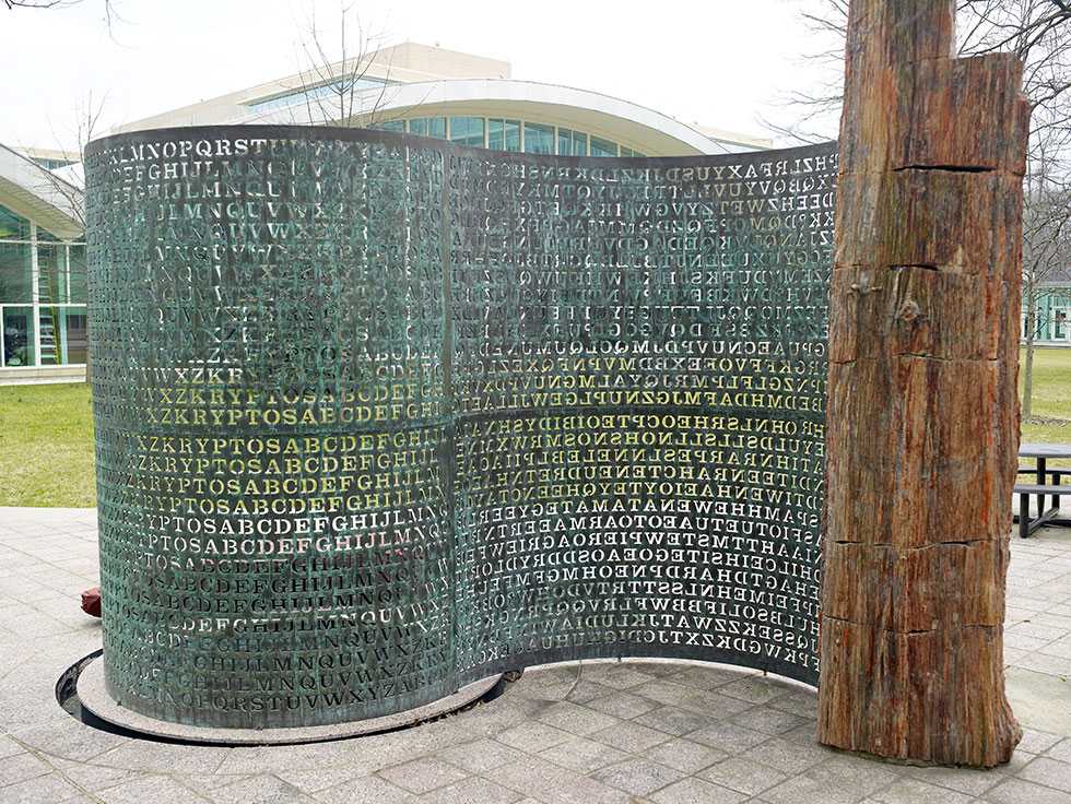 A full view of the "Kryptos" sculpture, with one side wrapping into an enclosed circle, then opening like a scroll, ending at a tall, distressed wooden mast.