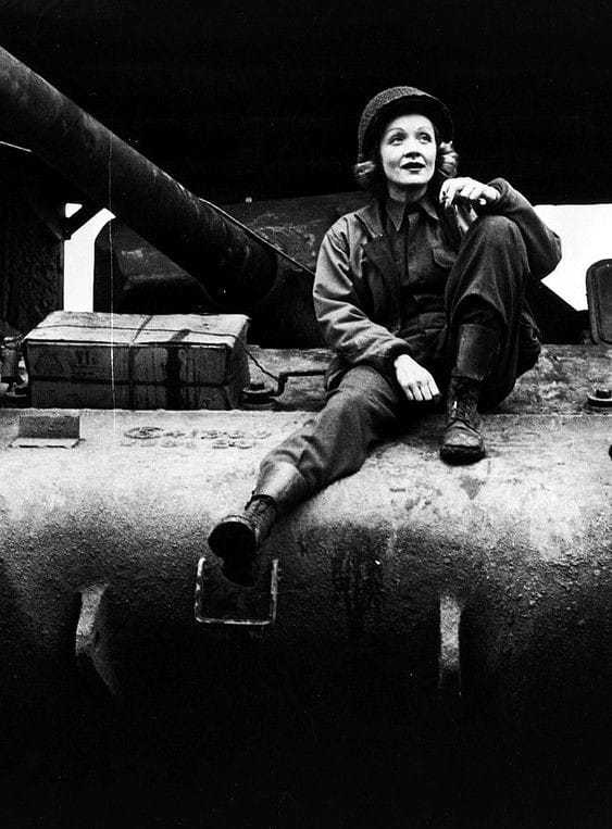 A black and white image of Marlene in uniform sitting on a crate, with one leg hanging of the end, looking upright towards the sky.