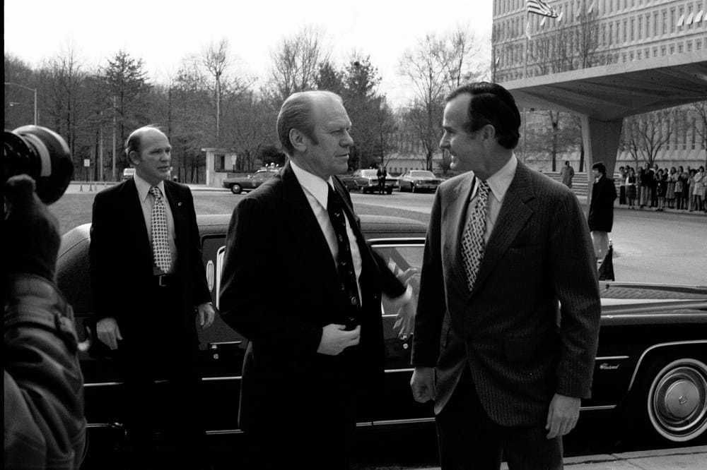 A black and white candid image of Bush and two men outside of the CIA Headquarters building standing in front of a car.