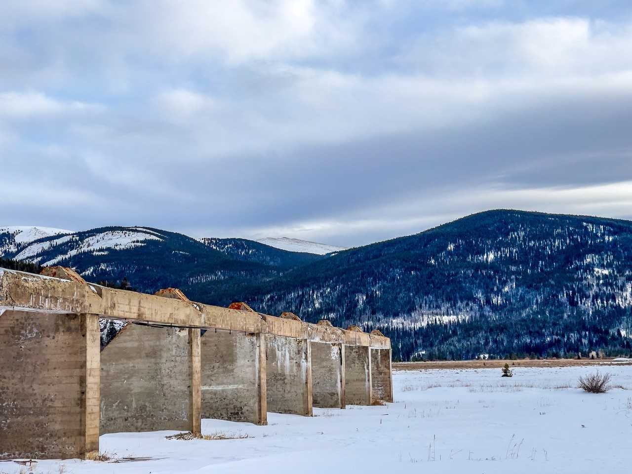 A snowy mountain scene with a an open plain in the center and an old concrete skeletal structure from WWII on the left side of the image.