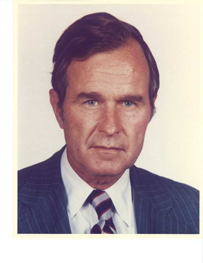 A headshot of George H. W. Bush in a blue suit with a blue and grey striped tie.