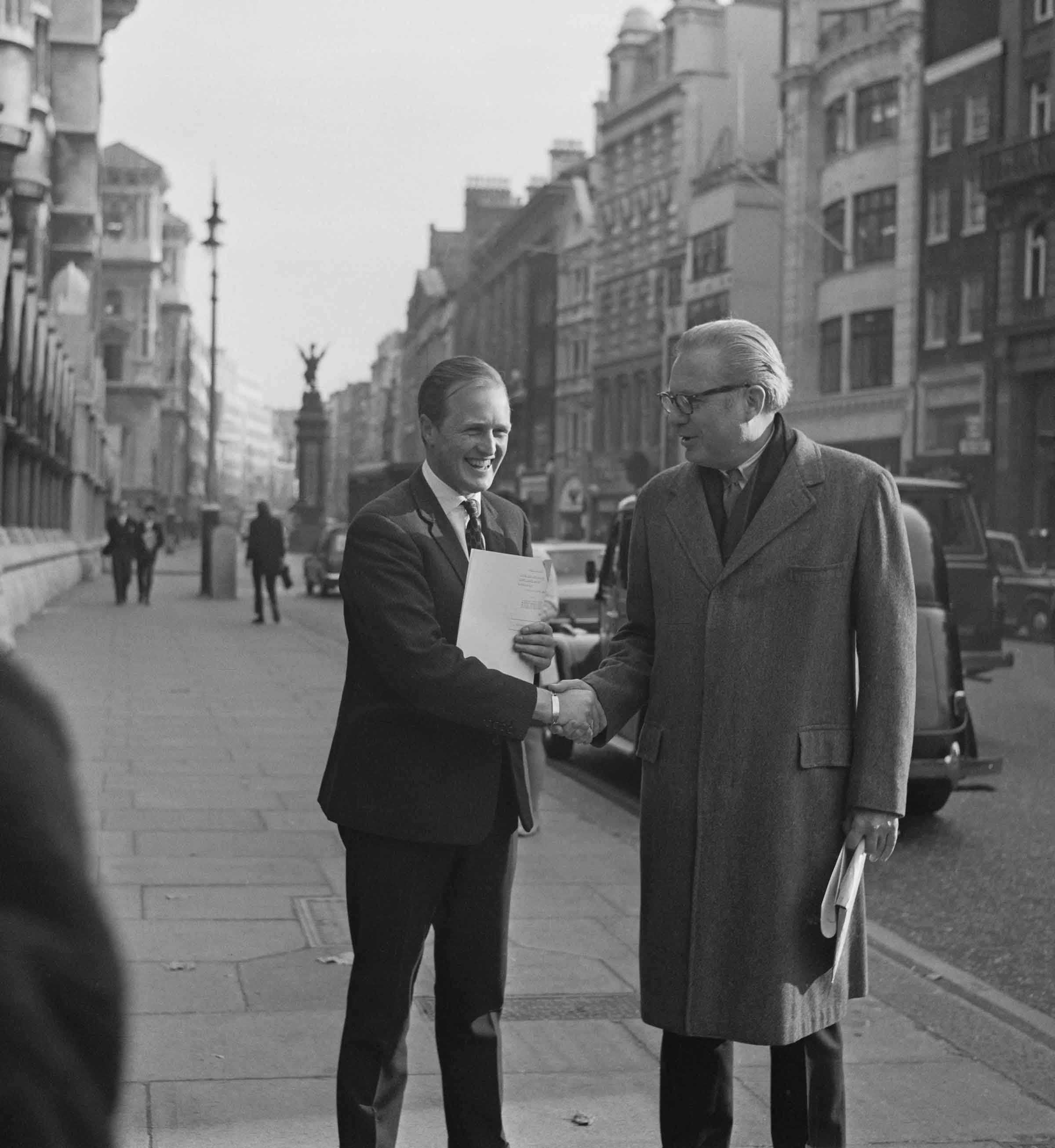 black and white photo of two men in the street shaking hands surrounded by tall buildings