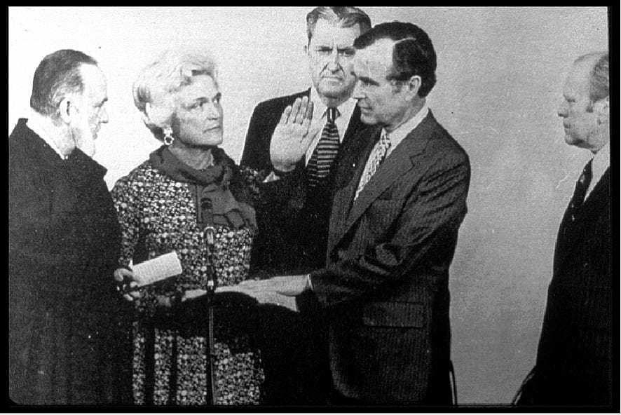 A black and white image of Bush surrounded by a woman and three men with his left hand placed on a book and his right hand raised in the air.