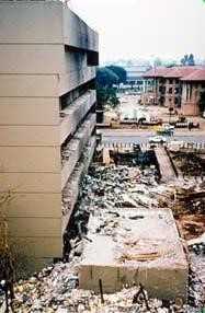 The aftermath of a bomb with debris where a building once stood and black ash covering the building next to the debris.