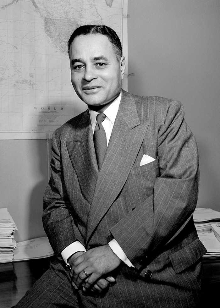 black and white photograph of man sitting on table with his hands crossed in his lap.