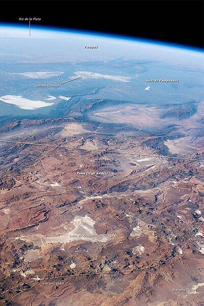 This panorama looking southeast across the South American continent was taken from the International Space Station almost directly over the Atacama Desert near Chile&apos;s Pacific coast. The high plains (3000-5000 m, 13,000-19,000 ft) of the Andes Mountains, also known as the Puna, appear in the foreground, with a line of young volcanoes (dashed line) facing the much lower Atacama Desert (1000-2000 m elevation). Several salt-crusted dry lakes (known as salars in Spanish) occupy the basins between major thrust faults in the Puna. Salar de Arizaro (foreground) is the largest of the dry lakes in this view. The Atlantic Ocean coastline, where Argentina&apos;s capital city of Buenos Aires sits along the R&#xed;o de la Plata, is dimly visible at image top left. Near image center, the transition (solid line) between two distinct geological zones, the Puna and the Sierras Pampeanas, creates a striking landscape contrast. Compared to the Puna, the Sierras Pampeanas mountains are lower in elevation and have fewer young volcanoes. Sharp-crested ridges are separated by wide, low valleys in this region. The Salinas Grandes - ephemeral shallow salt lakes - occupies one of these valleys. The general color change from reds and browns in the foreground to blues and greens in the upper part of the image reflects the major climatic regions: the deserts of the Atacama and Puna versus the grassy plains of central Argentina, where rainfall is sufficient to promote lush prairie grass, known locally as the pampas. The Salinas Grandes mark an intermediate, semiarid region. Image courtesy of NASA.