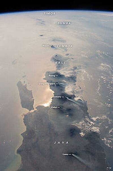 This photograph, taken from the International Space Station in 2015, looks eastward: Java is in the foreground, Bali and Lombok are near the center, and smaller islands trail off toward the horizon. The brightest reflection of the Sun off the sea surface silhouettes Surabaya, Indonesia’s second-largest city of almost 3 million. Against this background of glint as well as regional smoke, a line of volcanoes appears in sharp detail. Volcanoes are the backbone of the islands, which have been formed by the collision of the Australian tectonic plate (right) with the Asian plate (left). The name of each volcano appears in italics. White plumes - each about 80 km (50 mi) long - show that at least six volcanoes appeared to be emitting steam and smoke. The plumes are strikingly parallel, aligned with winds from the northeast. Image courtesy of NASA.