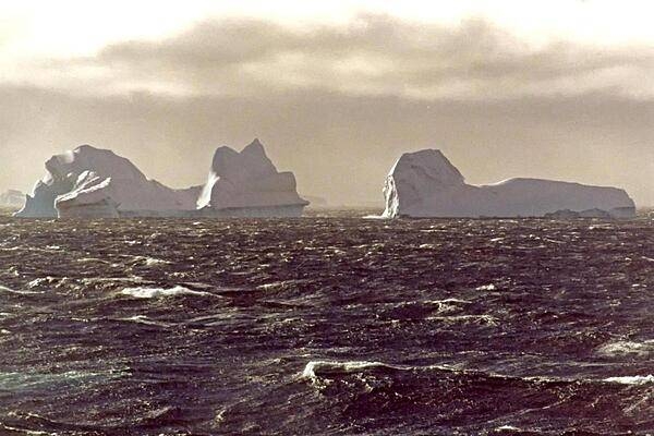 Looming icebergs cruise through rough, windswept seas. The icebergs (large pieces of fresh water ice) broke free from a snow-formed glacier or from the ice shelf that covers much of the continent.