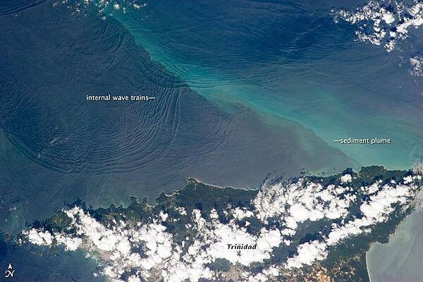 This space station photo of 18 January 2013, shows the north coast of Trinidad and a series of subtle, interacting arcs in the southeastern Caribbean Sea. These are known as &quot;internal waves,&quot; the surface manifestation of slow waves that move tens of meters beneath the sea surface. Internal waves produce enough of an effect on the sea surface to be seen from space, but only where they are enhanced due to reflection of sunlight, or sunglint, back towards the space station. 
The image shows at least three sets of internal waves interacting. The most prominent set (image top left) shows a packet of several waves moving from the northwest due to the tidal flow towards the north coast of Trinidad. Two less prominent, younger sets can be seen further out to sea. A very broad set enters the view from the north and northeast, and interacts at image top center with the first set. All the internal waves are probably caused by the shelf break near Tobago (outside the image to top right). The shelf break is the step between shallow seas (around continents and islands) and the deep ocean. It is the line at which tides usually start to generate internal waves. 
Over the island of Trinidad, the heating of the land surface sets off the growth of cumulus clouds. Off the coast, a light blue northwest-southeast trending plume at image center is sediment embedded in the Equatorial Current (also known as the Guyana Current). The current is transporting material to the northwest - in almost the opposite direction of the internal waves. The current flows strongly from east to west around Trinidad, all the way from equatorial Africa, driven by year-round easterly winds. Seafarers in the vicinity of Trinidad are warned that the current - and its local reverse eddies - make navigation complicated and sometimes dangerous for smaller craft in these waters.
Astronauts also have observed internal waves in other parts of the world, such as San Francisco and the Straits of Gibraltar. Photo courtesy of NASA.