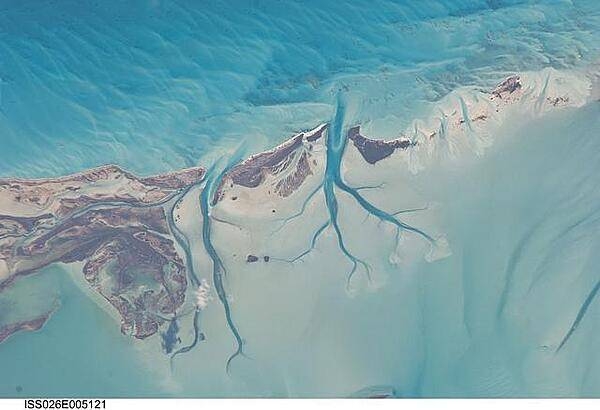 The islands of the Bahamas in the Caribbean Sea are situated on large depositional platforms (the Great and Little Bahama Banks) composed mainly of carbonate sediments ringed by fringing reefs - the islands themselves are only the parts of the platform currently exposed above sea level. The sediments are formed mostly from the skeletal remains of organisms settling to the sea floor; over geologic time, these sediments consolidate to form carbonate sedimentary rocks such as limestone. This detailed photograph taken from the International Space Station provides a view of tidal flats and tidal channels near Sandy Cay on the western side of Long Island, located along the eastern margin of the Great Bahama Bank. The continually exposed parts of the island have a brown coloration in the image, a result of soil formation and vegetation growth (left). To the north of Sandy Cay an off-white tidal flat composed of carbonate sediments is visible; light blue-green regions indicate shallow water on the tidal flat. Tidal flow of seawater is concentrated through gaps in the anchored land surface, leading to formation of relatively deep tidal channels that cut into the sediments of the tidal flat. The channels, and areas to the south of the island, have a vivid blue coloration that provides a clear indication of deeper water (center). Image courtesy of NASA.