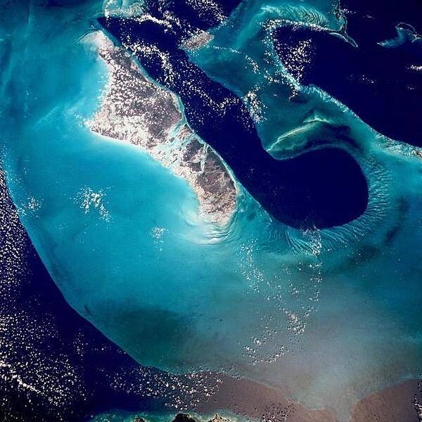 An even more stark view of the dark blue Tongue of the Ocean whose depths reach up to 1,800 m (6,000 ft). By comparison, the waters of the Bahama Platform (light blue) are frequently less than 15 m (50 ft) deep. They are warm and become extremely salty due to evaporation and limited circulation from the open ocean. Crystals of aragonite, a calcium carbonate mineral derived from the shells of single-celled marine organisms, and direct precipitation, form into oolites (small spherical grains of limestone) as the tidal currents swirl back and forth. Lithification of the carbonate sands produces an oolite limestone. Although the water is warm and clear, corals do not live in these shallows, probably because of the salt content. Though chemically very similar, the rocks resulting from this precipitating process have a quite different origin from those formed from coral reefs. Image courtesy of NASA.