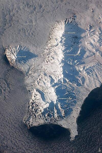 The Kuril Island chain is built from a line of volcanoes, an island arc, that extends from Russia&apos;s Kamchatka Peninsula to northern Japan. Island arcs form along an active boundary between two tectonic plates, with one being driven beneath the other (subduction). Magma generated by subduction feeds volcanoes - and eventually volcanic islands - over the subduction boundary. Paramushir Island in the northern Kurils is an example of a large island built by several volcanoes over geologic time. This astronaut photograph shows the southern end of Paramushir Island after a snowfall. The western slopes of the mountains are brightly illuminated, while the eastern slopes are in shadow.
Four major volcanic centers create this part of the island. Fuss Peak (image center left) is an isolated stratovolcano connected to the main island via an isthmus. Fuss Peak last erupted in 1854.
The southern tip of the island is occupied by the Karpinsky Group of three volcanic centers. A minor eruption of ash following an earthquake occurred on this part of the island in 1952. 
The Lomonosov Group to the northeast (image center) includes four cinder cones and a lava dome.
The most recent volcanic activity on Paramushir Island occurred in 2008 at the Chikurachki cone located along the northern coastline of the island at image top center. The summit of this volcano, 1,816 m (5,958 ft) above sea level, is the highest on Paramushir Island.
Much of the Sea of Okhotsk visible in the image is covered with low clouds that often form around the islands in the Kuril chain. The clouds are generated by moisture-laden air passing over the cool sea/ocean water, and they typically wrap around the volcanic islands. Image courtesy of NASA.