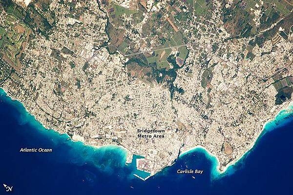 Bridgetown is the capital city of the island nation of Barbados, located to the east of the Lesser Antilles Island chain. While Barbados is considered part of the Lesser Antilles, it is located within the western Atlantic Ocean rather than the Caribbean Sea. Besides being the seat of government, Bridgetown is the largest city in Barbados. It and the surrounding towns that make up the Greater Bridgetown area are located along the southwestern coastline of the island. The metropolitan area is readily recognizable in this astronaut photograph due to the gray and white rooftops and street grids (image center) that contrast with green vegetated fields and riverside areas of the island's interior to the northeast (image top center). Bridgetown is a major port destination for both commercial and cruise ships serving the eastern Caribbean - several ships are visible within Carlisle Bay. Water color in the image changes from light blue along the coastline - indicating shallow water - to the dark blue of deeper water away from the island. Photo courtesy of NASA.