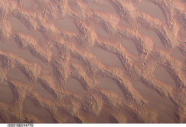 Photo shows classic large and small sand masses of the central Sahara Desert, where wind is a more powerful land-shaping agent than water. &quot;Draa&quot; dunes (from the Arabic for &quot;arm&quot;) are very large masses of sand, and they appear here as the broad network of yellow-orange sand masses, with smooth-floored, almost sand-free basins between them. These sand masses lie in the western part of Libya&apos;s vast Marzuq Sand Sea. Geologists think that the draa of the Marzuq were probably formed by winds different from the prevailing north-northeast winds of today. Numerous smaller dunes have developed on the backs of the draa. Three distinct dune types are visible: longitudinal dunes, which are more or less parallel with the north winds; transverse dunes, which are usually more curved and formed at right angles to the wind; and star dunes, in which several linear arms converge towards a single peak. Image courtesy of NASA.