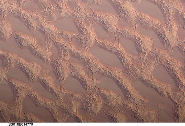 Photo shows classic large and small sand masses of the central Sahara Desert, where wind is a more powerful land-shaping agent than water. &quot;Draa&quot; dunes (from the Arabic for &quot;arm&quot;) are very large masses of sand, and they appear here as the broad network of yellow-orange sand masses, with smooth-floored, almost sand-free basins between them. These sand masses lie in the western part of Libya&apos;s vast Marzuq Sand Sea. Geologists think that the draa of the Marzuq were probably formed by winds different from the prevailing north-northeast winds of today. Numerous smaller dunes have developed on the backs of the draa. Three distinct dune types are visible: longitudinal dunes, which are more or less parallel with the north winds; transverse dunes, which are usually more curved and formed at right angles to the wind; and star dunes, in which several linear arms converge towards a single peak. Image courtesy of NASA.