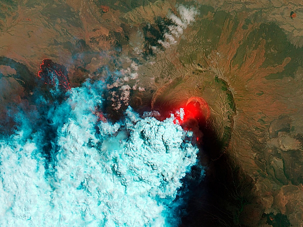 Satellite view of the Nabro Volcano taken on 24 June 2011. The bright red areas indicate hot surfaces. Hot volcanic ash glows above the vent, located in the center of Nabro’s caldera. To the west of the vent, portions of an active lava flow (particularly the front of the flow) are also hot. The speckled pattern on upstream portions of the flow are likely due to the cool, hardened crust splitting and exposing fluid lava as the flow advances. The bulbous blue-white cloud near the vent is likely composed largely of escaping water vapor that condensed as the plume rose and cooled. The whispy, cyan clouds above the lava flow are evidence of degassing from the lava. Image courtesy of NASA.