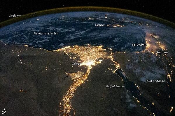 One of the fascinating aspects of viewing Earth at night is how well the lights show the distribution of people. In this view of Egypt, we see a population almost completely concentrated along the Nile Valley, just a small percentage of the country&apos;s land area. 
The Nile River and its delta look like a brilliant, long-stemmed flower in this astronaut photograph of the southeastern Mediterranean Sea. The Cairo metropolitan area forms a particularly bright base of the flower. The smaller cities and towns within the Nile Delta tend to be hard to see amidst the dense agricultural vegetation during the day. However, these settled areas and the connecting roads between them become clearly visible at night. Likewise, infrastructure and urbanized regions along the Nile River also become apparent.
Another brightly lit region is visible along the eastern coastline of the Mediterranean, the Tel-Aviv metropolitan area in Israel (image right). To the east of Tel-Aviv lies Amman, Jordan. The two major water bodies that define the western and eastern coastlines of the Sinai Peninsula, the Gulf of Suez and the Gulf of Aqaba, are outlined by lights along their coastlines (image lower right). The city lights of Paphos, Limassol, Larnaca, and Nicosia are visible on the island of Cyprus (image top).
Scattered blue-grey clouds cover the Mediterranean Sea and the Sinai, while much of northeastern Africa is cloud-free. The thin yellow-brown band tracing the Earth&apos;s curvature at image top is airglow, a faint band of light emission that results from the interaction of atmospheric atoms and molecules with solar radiation at approximately 100 km (60 mi) altitude. Photo courtesy of NASA.