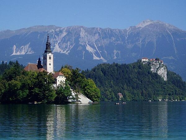 Lake Bled in Slovenia, with the Church of the Assumption at the left on little Bled Island, Bled Castle atop the cliffs on the right, and the Julian Alps towering in the background. Small boats with strong oarsmen ferry visitors to the island - to the musical accompaniment of an accordion.