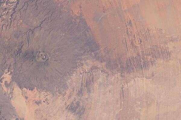 Emi Koussi Volcano and Aorounga Impact Crater in Chad are featured in this striking image photographed from the International Space Station. The two circular landscape features show craters produced by very different geological processes. At left, the broad grey-green shield volcano of Emi Koussi is visible (its 3,415 m summit is the highest in the Sahara region). The circular Aorounga Impact Crater is located approximately 110 km to the southeast of Emi Koussi and has its origin in forces from above rather than eruptions from below. The Aorounga structure is thought to record a meteor impact approximately 345-370 million years ago. Image courtesy of NASA.