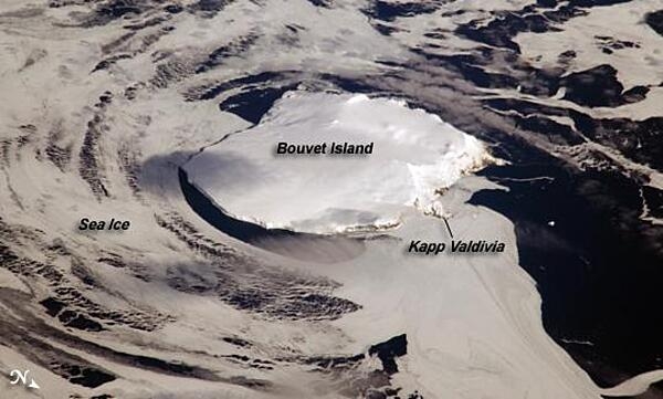 Bouvet Island is one of the most remote islands in the world; Antarctica, over 1,600 km (1,000 mi) to the south, is the nearest land mass. Located near the junction of three tectonic plates, the island is mostly formed from a shield volcano that is almost entirely covered by glaciers. The prominent Kapp (Cape) Valdivia on the northern coastline is a peninsula formed by a lava dome. It is only along the steep cliffs of the coastline that the underlying dark volcanic rock is visible against the white snow and ice blanketing the island. Photo courtesy of NASA.