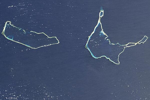 Majuro and Arno Atolls appear as meandering lines of delicate white against a darker ocean background in this satellite image. Areas of shallow water near the shore appear iridescent blue, especially in the southwest portion of each atoll and off the northwest tip of Arno Atoll. Overhead, tiny white clouds dot the sky. Majuro Atoll is 40 km (25 mi) in circumference, and it partially encloses a lagoon of nearly 300 sq km (115 sq mi). The lagoon is far bigger than the atoll itself, whose land area is less than 10 sq km (4 sq mi); walking from the lagoon side to the ocean side of the atoll takes only minutes. Neighboring Arno Atoll encloses three lagoons: two &quot;pinched lagoons&quot; in the northeast and northwest, and a main lagoon of nearly 340 sq km (130 sq mi). Arno&apos;s total land area is just 13 sq km (5 sq mi). Scientists have long understood that coral atolls develop around volcanic islands and remain there after those islands have eroded away. To what extent the final shape of an atoll is influenced by wind and ocean wave activity, however, remains a topic of debate. Image courtesy of NASA.