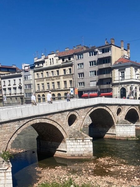 The Latin Bridge is the oldest of over a dozen bridges crossing the Miljacka River in Sarajevo. The original wooden bridge dates to around 1541 but was replaced by a stone structure in 1565.  A 1791 flood damaged the bridge, and it was rebuilt around 1798. Constructed while Bosnia was under the Ottoman Empire, the bridge acquired its name from the fact that it connected with the Catholic quarter of the city (“Latinluk”) on the right bank of the river. The stone and gypsum bridge has four arches and sits on three pillars that have two relieving openings or “eyes” above them. The distinctive “eyes” also appear on the Sarajevo coat of arms. The Latin Bridge acquired a place in history as the spot where World War I began.  On 28 June 1914 at the foot of the bridge, a Serb nationalist, Gavrilo Princip, shot and killed Franz Ferdinand, heir presumptive to the Austro-Hungarian throne, and his wife Sophie sparking  WWI.